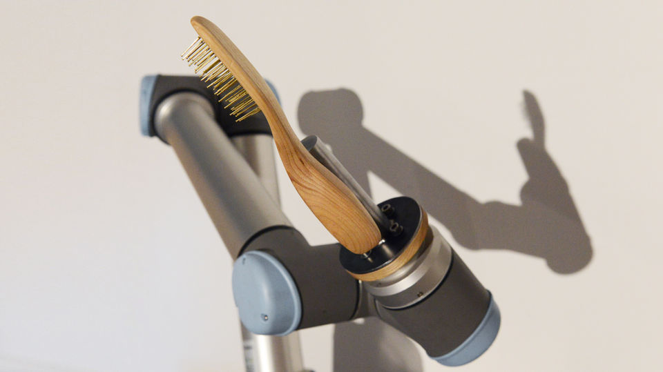 UR10 robotic arm holding a wooden hairbrush against a soft white wall, casting a human-shaped shadow.